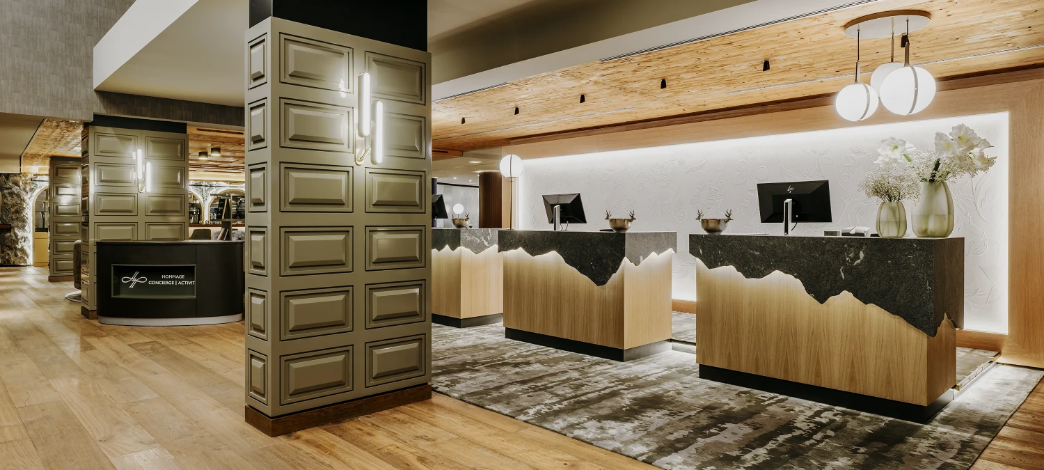 Dieses Foto zeigt den luxuri&ouml;sen Innenraum des Grand Tirolia Kitzb&uuml;hl Hotels. Man kann eine luxuri&ouml;se Lobby mit einer modernen Rezeption, bequemen Sitzm&ouml;beln, einem h&uuml;bschen Blumenarrangement und einer riesigen Glasfront erkennen.